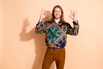 Trendy young man with long hair in patterned shirt showcasing hand gestures on a beige background