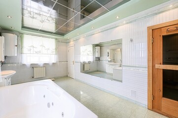 Bright bathroom with white tile, a large mirror, a tub, a window with floral curtains, and a sauna door. The ceiling has a reflective surface