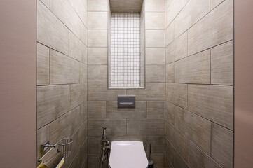 modern, tiled bathroom. The walls are covered in light brown, wood-look tiles, with a rectangular niche filled with white mosaic tiles. A toilet is centered at the bottom