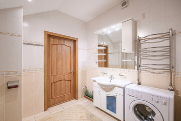 well-lit bathroom features a wooden door, tiled walls, a sink with a cabinet, a mirror with storage, a towel warmer, and a washing machine