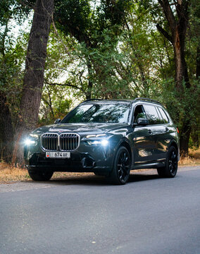 Bishkek, Kyrgyzstan - October 12, 2024: A dark gray 2024 BMW X7 is stationary on a paved road, bordered by dense green trees and foliage under natural light