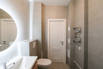 modern bathroom featuring grey tiles, a round mirror with a light, a white door, and fixtures