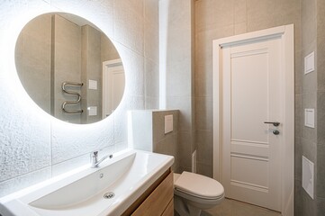 modern bathroom with grey tiles, a round illuminated mirror, a white sink on a wooden cabinet, and...