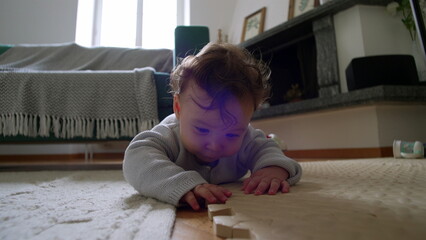 Baby lying on the floor, closely examining a wooden block, leaning down with curiosity, actively...