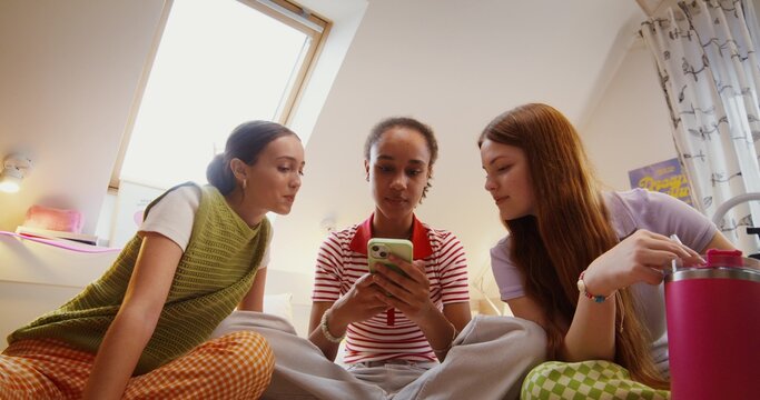 Three teenage girls are using a mobile phone and talking. They're sitting on the bed.