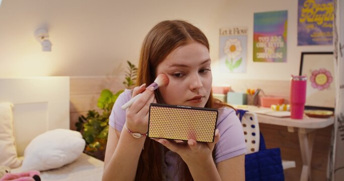 A girl does her makeup while sitting on the bed. She puts blush on her face with a brush.