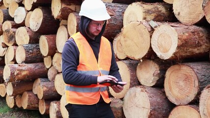 A worker in a safety vest and helmet is focused on a tablet in front of stacked logs at a lumber yard. He appears to be managing tasks and monitoring inventory in a forested setting - Powered by Adobe
