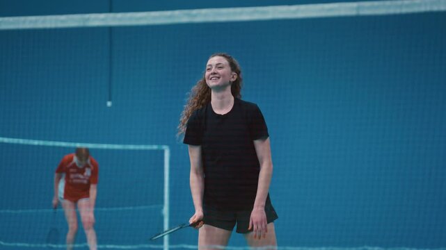 Portrait: red-haired smiling girl playing badminton indoors. Happy female athlete hits shuttlecock with a racket across a net. Practicing serve before playing in badminton on inside court