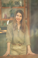 Smiling Light-Skinned European Woman in Olive Shirt Seen Through Window in Cozy Rustic Kitchen