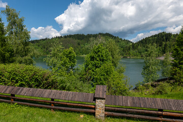 Fototapeta premium View of Czorsztyn Lake in Poland from behind a wooden fence. Green trees, calm water and forested hills under a partly cloudy sky in the background.
