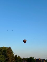 Colorful hot air balloon floats peacefully over rooftops and treetops at sunrise, with birds soaring in the clear blue sky portraing a tranquil moment in the countryside.