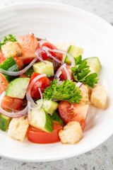 Fresh vegetable salad with tomatoes, cucumbers, red onions, parsley, croutons, and olives in white bowl on a speckled gray surface. vibrant colors and healthy ingredients make it appealing photography