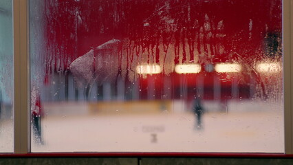 Foggy glass with ice rink view, blurred figures in background, condensation on window, winter indoor sports scene, hockey arena atmosphere, cold weather details