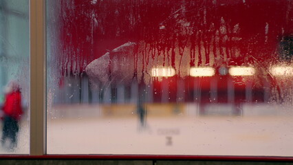 Foggy glass with ice rink view, blurred figures in background, condensation on window, winter indoor sports scene, hockey arena atmosphere, cold weather details