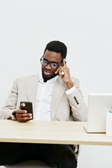 Confident smiling man of African descent wearing glasses and beige blazer sitting at desk with...