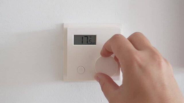 Hand touching smart thermostat, adjusting digital temperature settings, to increase the temperature, highlighting sleek home automation tech against minimalist white wall background