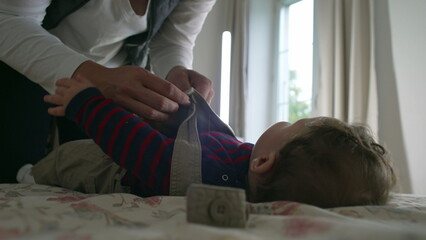 Mother fastening baby’s overalls, close-up hands adjusting straps while baby lies on the bed, calm interaction in cozy room with a mother and baby in morning routine