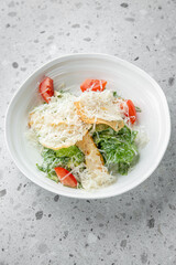 Fresh Caesar salad with grilled chicken, cherry tomatoes, romaine lettuce, croutons, and Parmesan cheese in white bowl on speckled gray countertop. Healthy and delicious meal with vibrant ingredients
