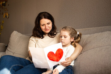 Little daughter with golden hair presents hand-drawn card to her smiling mother. Genuine moment of...