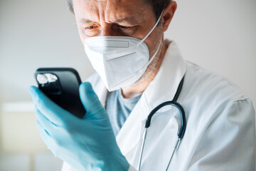 Tired exhausted male doctor wearing protective face mask using smartphone in hospital clinic office