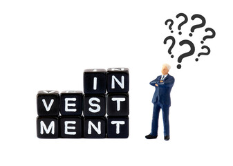 A miniature businessman figurine stands thoughtfully beside a stack of black letter blocks spelling "INVESTMENT" on a white background. Question marks float above his head, symbolizing financial uncer