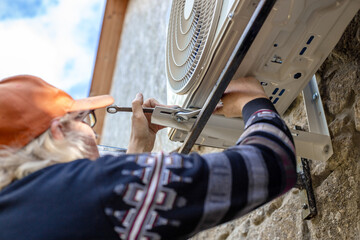 Obraz premium A man installs an outdoor unit of an air conditioner on iron mounting brackets, securing it with bolts and a wrench on the wall of a stone farmhouse. DIY installation