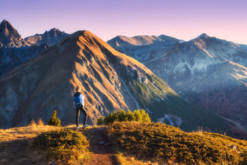 Young woman with backpack standing on grassy mountain trail in valley with high mountain peaks during golden hour sunset in autumn. Hiking and trekking in Italy, Livigno. Girl in Alps in fall