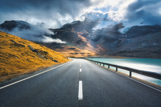 Empty asphalt road winding through a breathtaking mountain landscape, with majestic snowy peak with glacier and lake under dramatic cloudy sky at sunset. Road on mountain pass in swiss alps in autumn