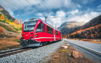 Red modern passenger train is moving on railway in swiss alps at sunset in autumn. Landscape with train in high mountains, road, orange trees in fall. Bernina Express, Switzerland. Mauntain pass