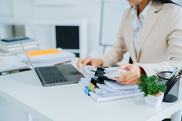 A young Asian businesswoman works alone at a desk with a laptop, stacks of papers and documents....