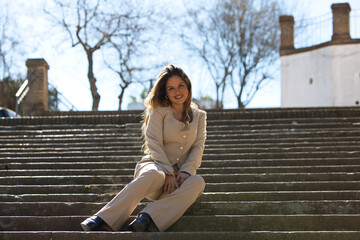 Blonde, young and beautiful woman sitting on the steps of the stairs in a street. The woman is...