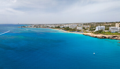 Aerial view of Ayia Napa, Cyprus, showing turquoise waters, sandy beaches, luxury resorts, white buildings, rocky outcrops, and green vegetation.