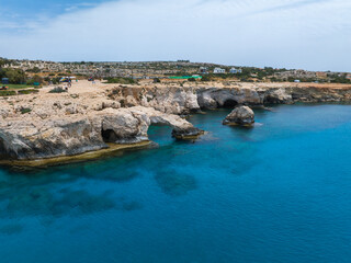 Limestone cliffs and natural rock arches extend into turquoise Mediterranean waters in Ayia Napa, Cyprus, under a clear sky with light clouds.