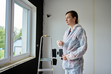 Woman in protective coveralls standing in front of unpainted wall with paint samples. Person...