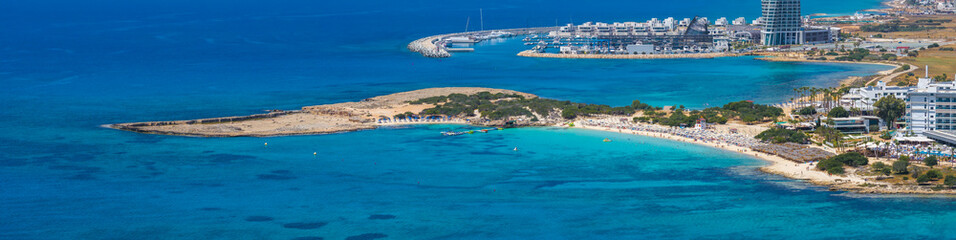 Aerial view of Nissi Beach in Ayia Napa, Cyprus, with turquoise waters, sandy shores, luxury hotels, a marina, yachts, and a tall modern building.