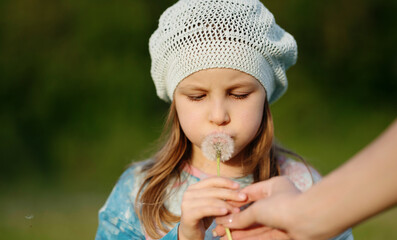 Little girl about to blow a dandelion with an adult hand helping her hold it. Sweet summer moment in a meadow. Close-up with natural light and soft focus.