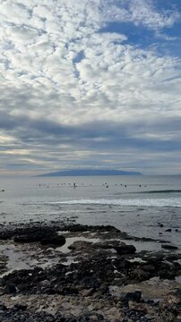 Vertical long-shot photo with wide-angle focal length, showing surfers in ocean waves near rocky shore, distant mountain, cloudy sky during daylight, at seaside leisure location