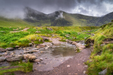 A serene and tranquil Carrauntoohil mountain scene unfolds, featuring a gently flowing stream surrounded by lush, vibrant greenery and dramatic clouds rolling overhead in the sky, Kerry, Ireland