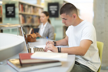 Young student guy reading books and working on laptop in library