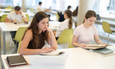 While working in library, girls student sit at table, read books, collect and structure information, masters new knowledge. Extracurricular reading, auxiliary literature, preparation for final test