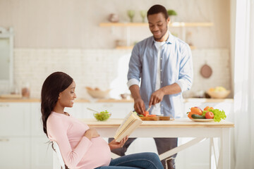 Black expectant lady reading book while her boyfriend cooking balanced breakfast in kitchen. Handsome African American guy making yummy vegetable salad for his pregnant girlfriend at home