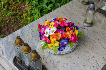 Colorful artificial flowers arranged in bowl with memorial candles on cemetery grave marker