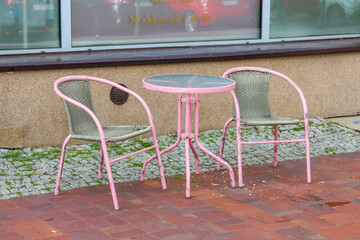A small outdoor cafe setting with two pink chairs and a round table on a paved surface with red...
