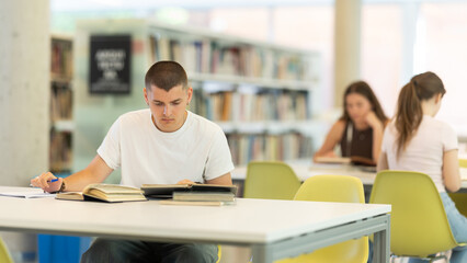 Fototapeta premium European guy is sitting at a table, reading a book and making notes in a notebook, he is preparing to enter the university in the new academic year. Students study in the library