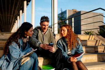 Three cheerful university students are sitting on campus steps, sharing content on a smartphone and enjoying the sunny day