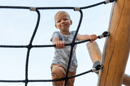 Happy blond boy climbing rope net at playground on sunny day, smiling and enjoying outdoor activity. Concept of childhood adventure, playtime and physical development. High quality photo