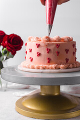 Red hearts being piped on Pink buttercream cake on a cake plate, Cake covered in pink frosting, process of decorating a cake with red buttercream hearts