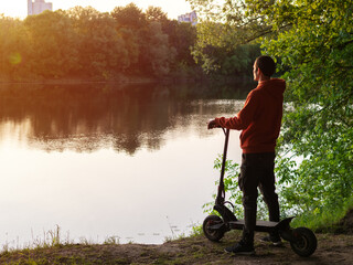 A man on an electric scooter stands by the river and enjoys nature at sunset.