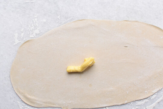 Overhead view of butter being spread on rolled out paratha dough, top view of flaky flatbread dough being covered with ghee, process of making paratha	