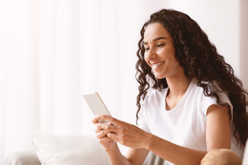 Fototapeta premium Young pretty woman in white t-shirt using modern smartphone and smiling, checking social media or checking with her friends, sitting next to window at home, closeup, copy space